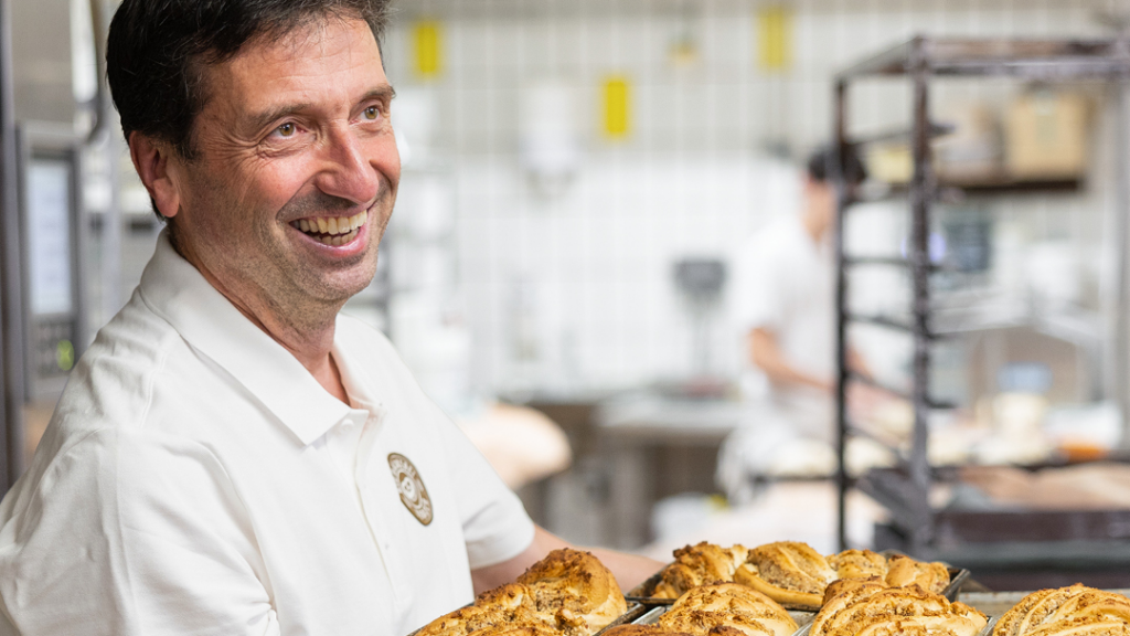 Ein lächelnder Bäcker in weißem Poloshirt hält in einer Backstube ein Blech mit frisch gebackenen, goldbraunen Broten in den Händen, während im Hintergrund weitere Backwarenregale und eine unscharfe arbeitende Person zu sehen sind.