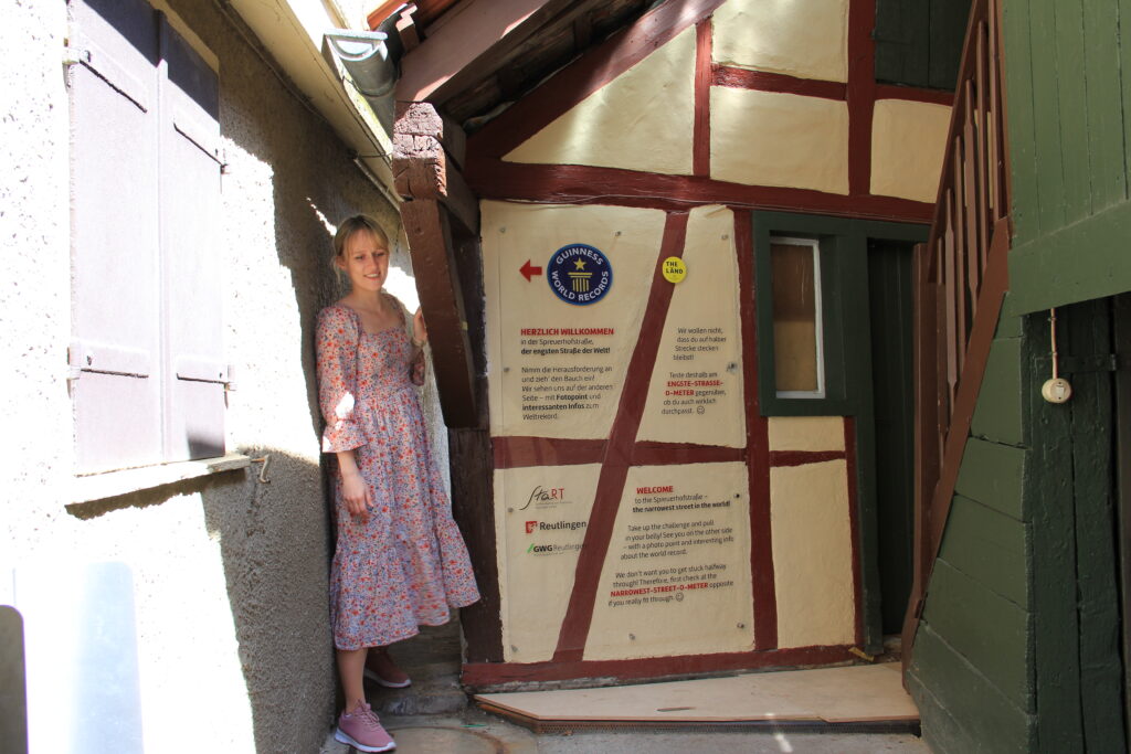 Eine junge Frau steht l&auml;chelnd in einer sehr schmalen Gasse zwischen Fachwerkh&auml;usern, neben einer Tafel mit Informationen zur &bdquo;engsten Stra&szlig;e der Welt&ldquo;.