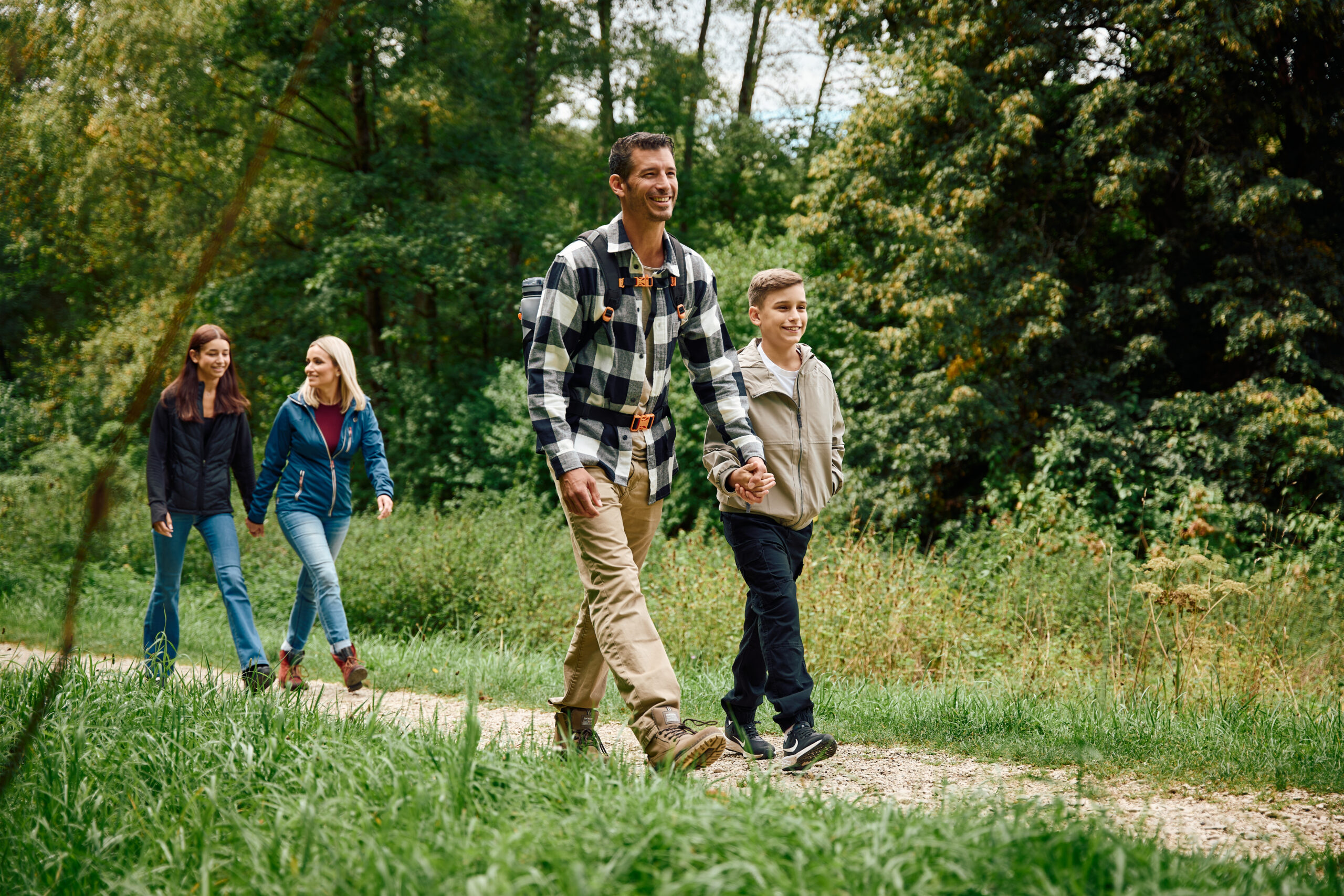 Vier Personen &ndash; zwei Erwachsene und zwei Kinder &ndash; gehen gemeinsam auf einem schmalen Waldweg spazieren. Im Vordergrund l&auml;uft ein Mann in kariertem Hemd und beige Hose, der einen Jungen an der Hand h&auml;lt. Dahinter gehen zwei Frauen nebeneinander; eine tr&auml;gt eine blaue Jacke, die andere eine schwarze Jacke. Alle wirken entspannt und fr&ouml;hlich. Um sie herum ist &uuml;ppiges Gr&uuml;n mit hohen B&auml;umen und Gras, und der Weg ist von Natur umgeben.