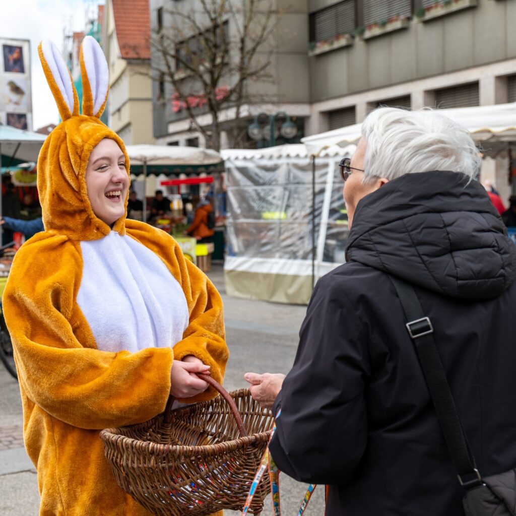 Osterhase im Kostüm verteilt lachend Süßigkeiten aus einem Korb an eine ältere Frau auf dem Marktplatz in der Reutlinger Innenstadt.