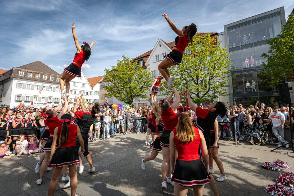 Eine Cheerleading-Gruppe in rot-schwarzen Uniformen zeigt auf einem belebten Platz vor Publikum eine Wurfakrobatik, bei der mehrere Sportlerinnen durch die Luft fliegen. Im Hintergrund stehen Zuschauerinnen und Zuschauer zwischen B&auml;umen und Geb&auml;uden unter blauem Himmel.