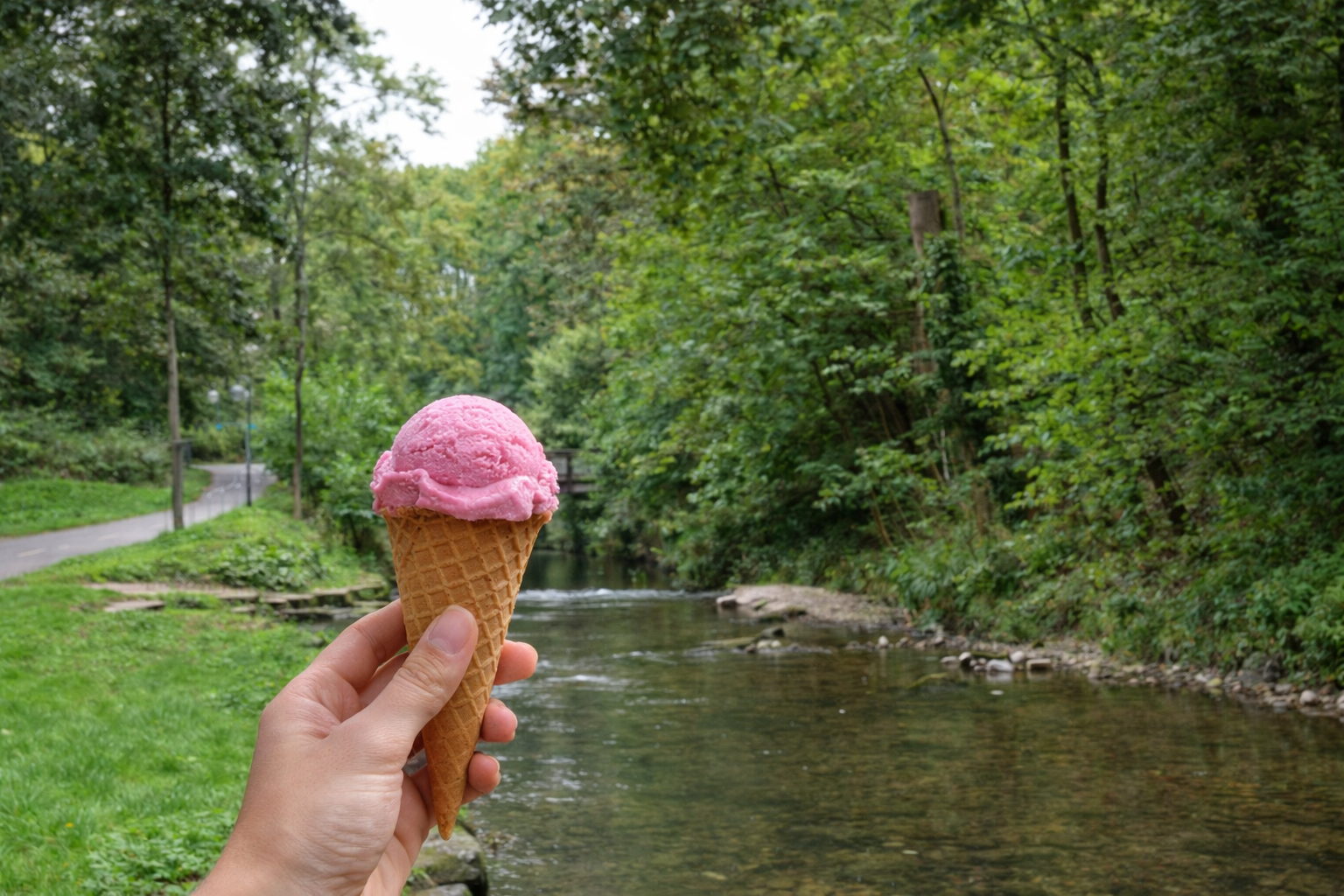 Hand mit rosa Eis vor einem schmalen Fluss in gr&uuml;ner, baumbestandener Umgebung.