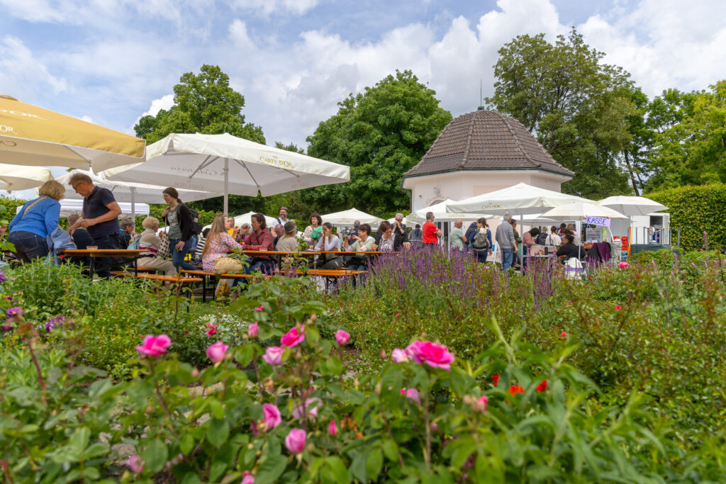 Belebter Garten mit G&auml;sten an Biertischen, Sonnenschirmen, Blumenbeeten und kleinem Geb&auml;ude im Hintergrund.