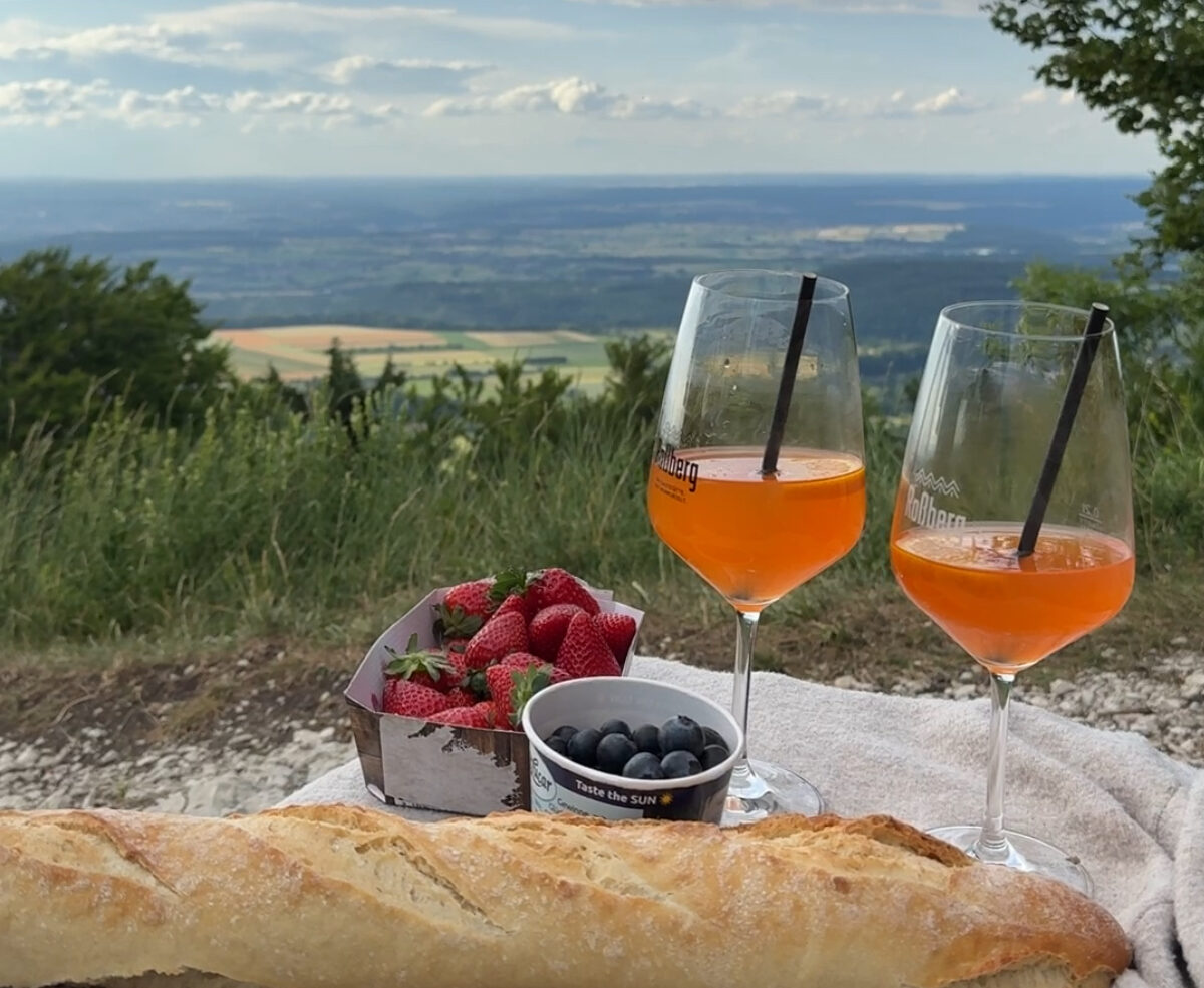 Gem&uuml;tliches Picknick mit Baguette, Beeren und zwei Drinks auf einer Decke mit weitem Panoramablick &uuml;ber die Landschaft.