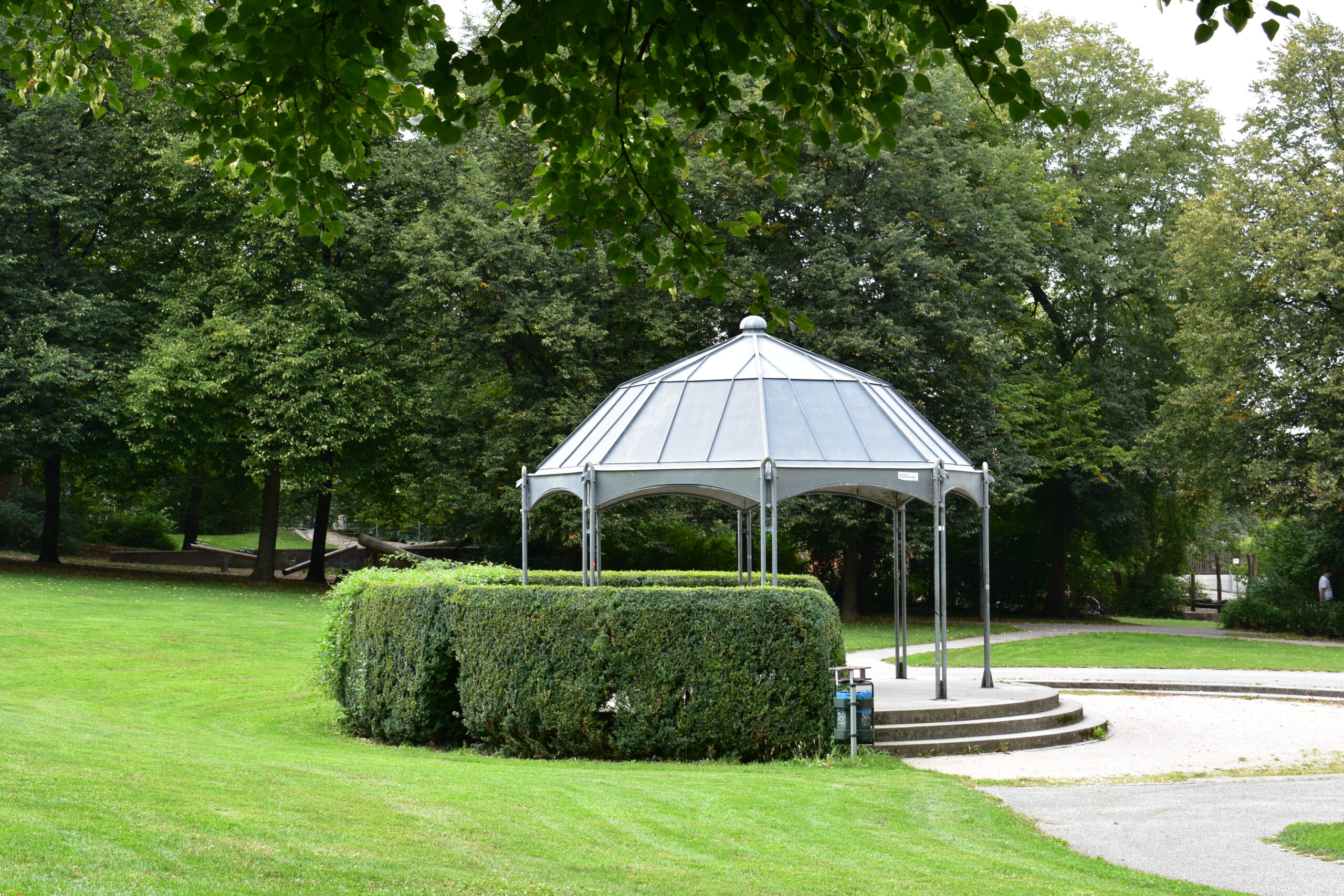 Heller Pavillon in einer gr&uuml;nen Parkanlage mit Rasen, B&auml;umen und niedriger Hecke.