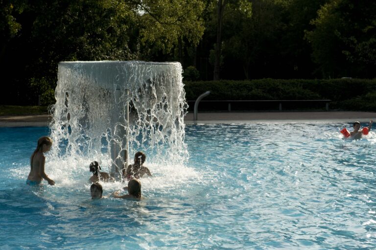 Mehrere Kinder spielen fr&ouml;hlich in einem Freibadbecken direkt unter einem gro&szlig;en Wasserpilz-Brunnen, w&auml;hrend ein weiteres Kind mit Schwimmfl&uuml;geln im Hintergrund schwimmt.