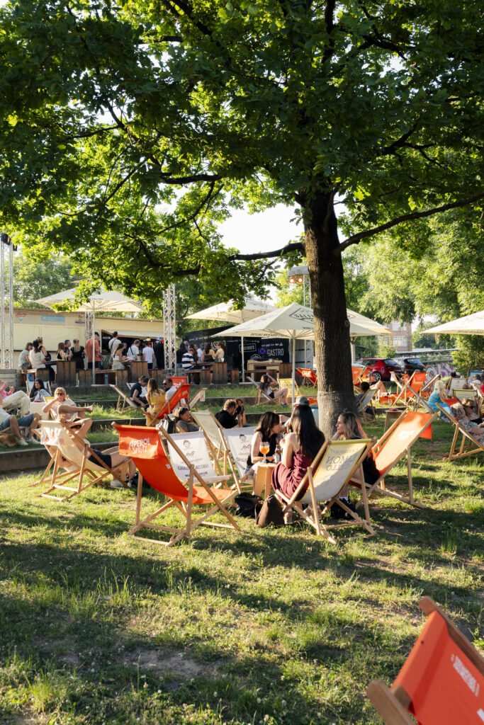 Eine sommerliche Open-Air-Gastronomie im Gr&uuml;nen unter einem gro&szlig;en, schattigen Baum. Auf einer Rasenfl&auml;che im Vordergrund stehen gem&uuml;tliche Liegest&uuml;hle in Orange und Wei&szlig;, in denen G&auml;ste entspannen und Drinks genie&szlig;en. Im Hintergrund herrscht reges Treiben an einer h&ouml;lzernen Bar und an Foodtrucks unter wei&szlig;en Sonnenschirmen. Das Sonnenlicht f&auml;llt warm durch die Bl&auml;tter des Baumes und sorgt f&uuml;r eine lebendige Afterwork- und Biergarten-Atmosph&auml;re.