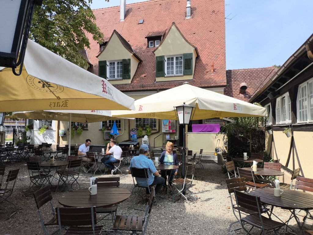 Sonnige Au&szlig;enterrasse eines gem&uuml;tlichen Restaurants mit gro&szlig;en Sonnenschirmen, Holztischen und G&auml;sten auf einem Kiesplatz vor einem traditionellen Geb&auml;ude mit rotem Ziegeldach.