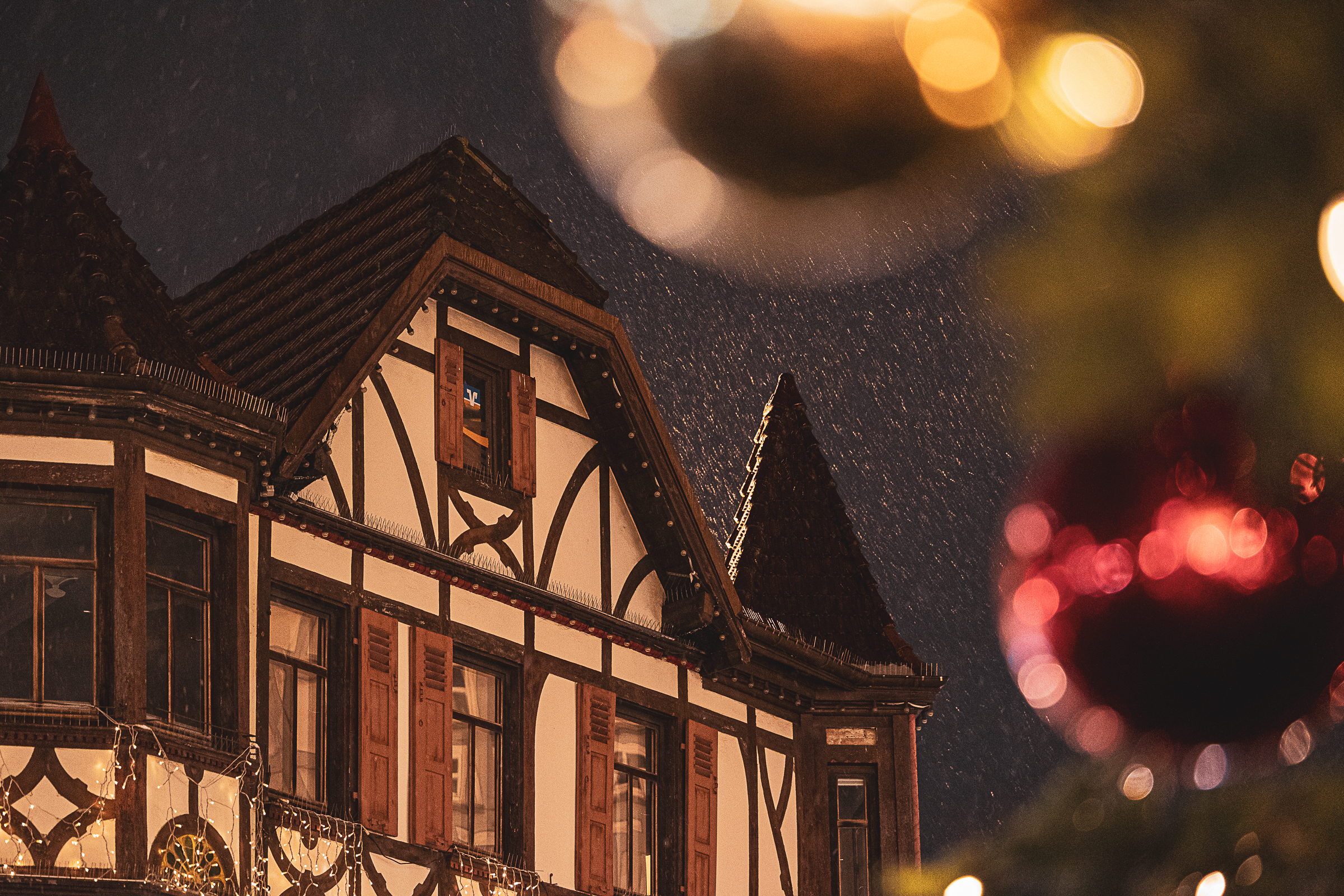 Nächtliche Aufnahme bei dichtem Schneetreiben in einer Stadt. Links ist die beleuchtete, weiße Fassade eines Wohnhauses mit mehreren Fenstern zu sehen. Im Hintergrund ragt die beleuchtete, steinerne Spitze eines gotischen Kirchenturms in den dunklen Nachthimmel.