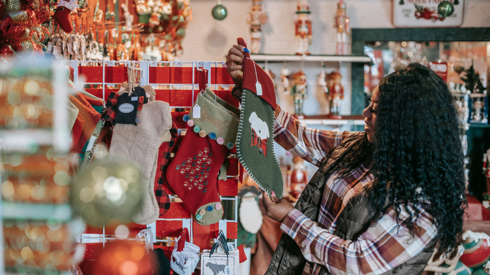 Ein stimmungsvolles Foto aus einem Weihnachtsgeschäft, das eine Frau zeigt, die einen bestickten grünen Weihnachtsstrumpf begutachtet. Im Hintergrund sind Regale voller Nussknacker und anderer festlicher Dekorationen zu sehen, sowie Unmengen von roten und goldenen Ornamenten im Vordergrund.