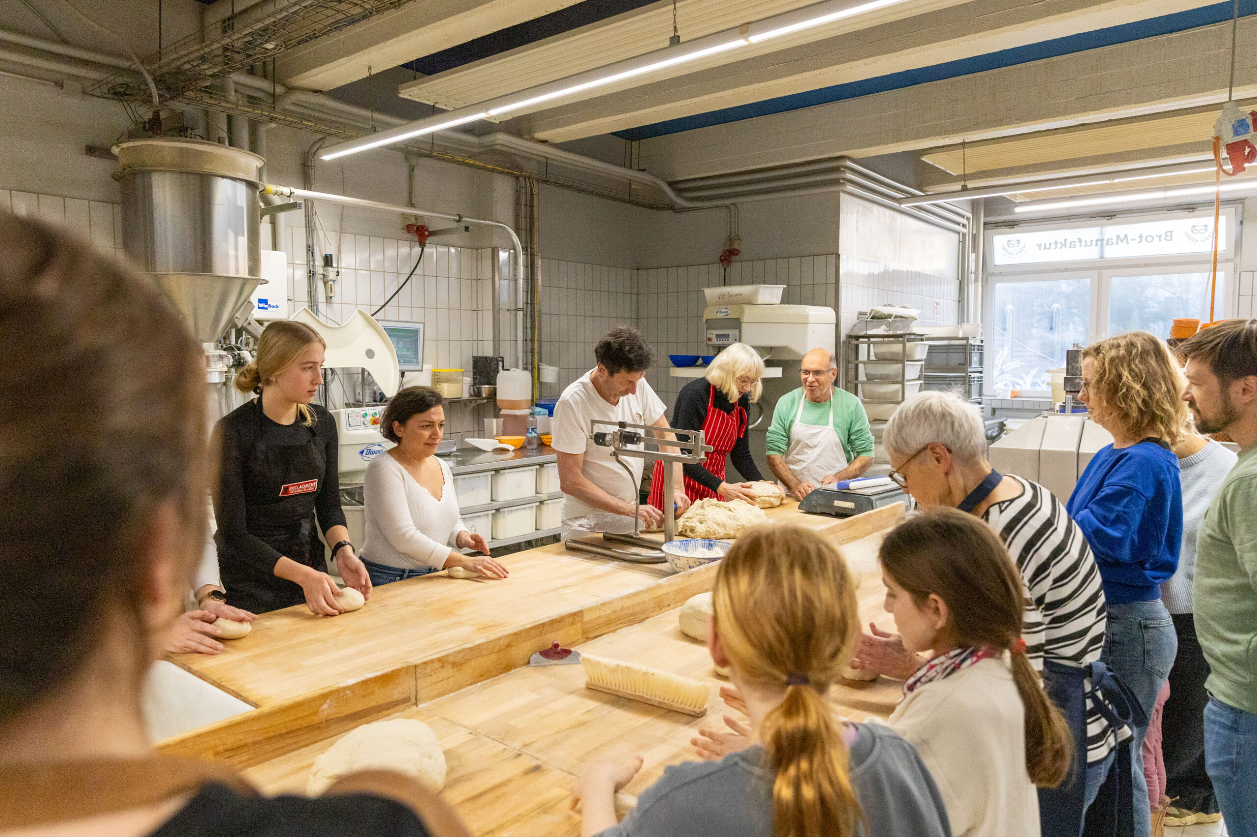 Ein Backworkshop in einer Backstube: Erwachsene und Kinder kneten gemeinsam Teig an gro&szlig;en Holztischen.