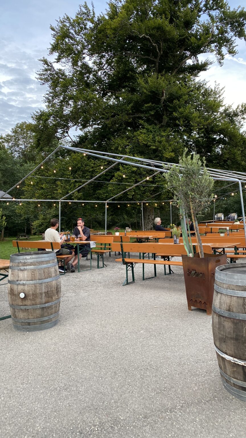 Au&szlig;enbereich einer Gastronomie mit Bierzeltgarnituren unter einem offenen Zeltgestell mit Lichterkette, flankiert von Holzf&auml;ssern und B&auml;umen.