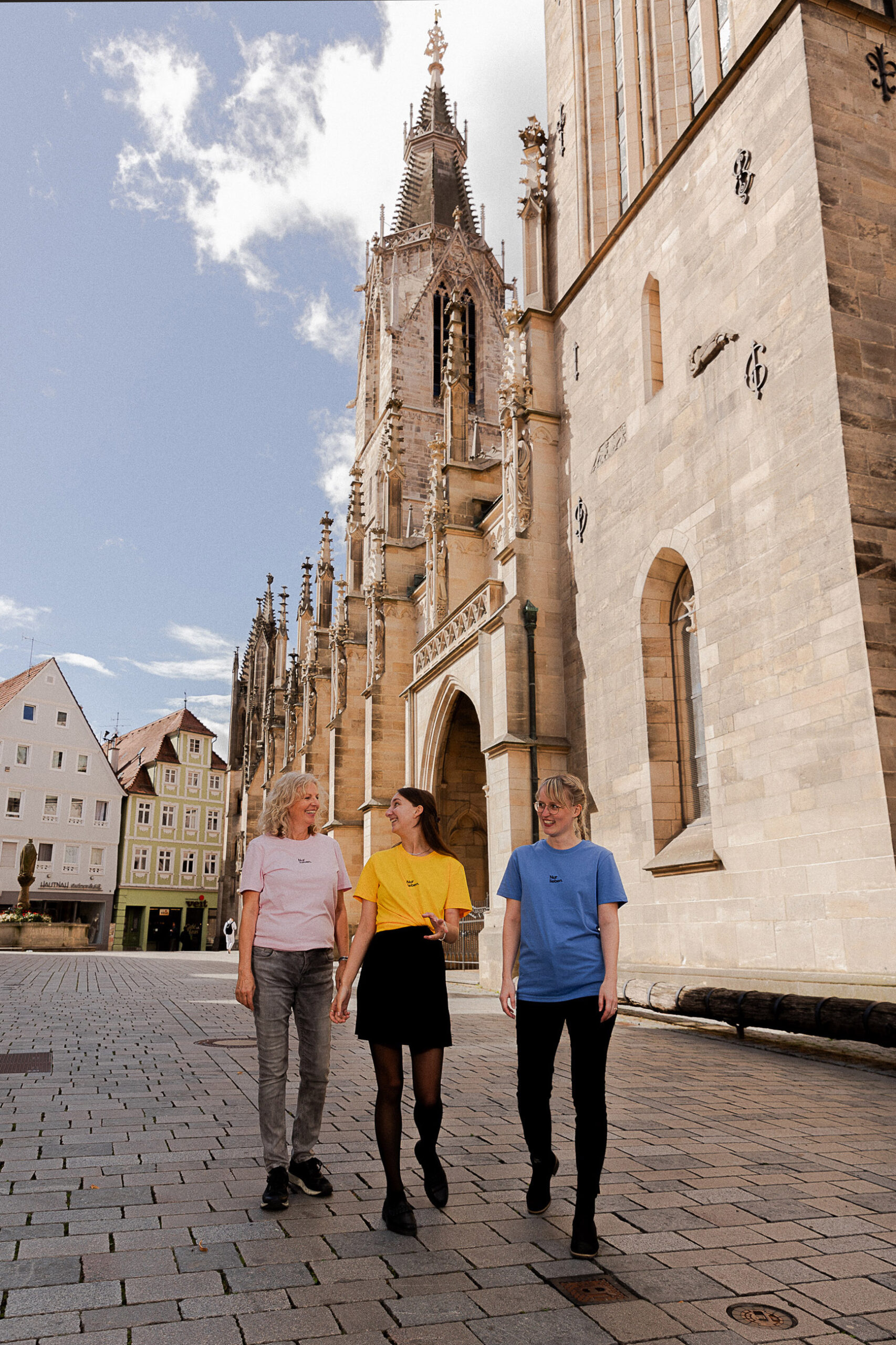 Drei Frauen in bunten T-Shirts gehen lachend über einen gepflasterten Platz vor einer großen gotischen Kirche.