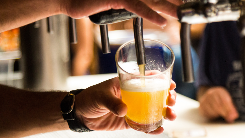 Nahaufnahme: Hand h&auml;lt ein Glas unter einen Zapfhahn, aus dem frisches, goldenes Bier mit Schaumkrone gezapft wird.
