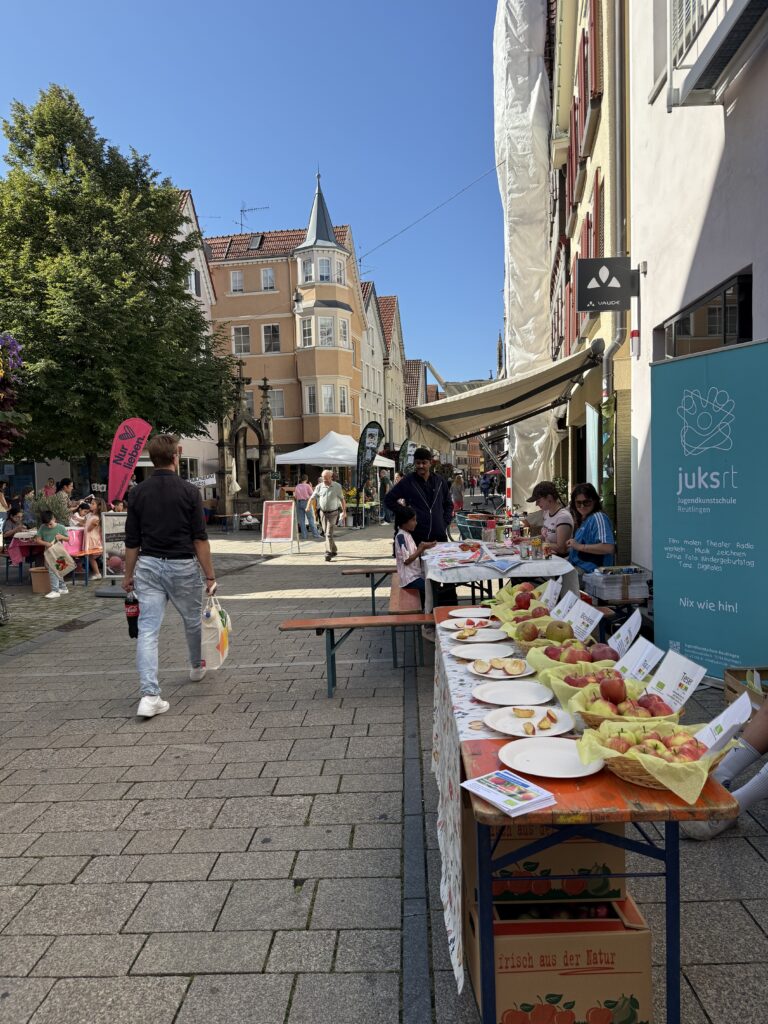 Eine Fotografie einer sonnigen, belebten Szene in der Altstadt von Reutlingen. Im Vordergrund ist ein Tisch mit Äpfeln drauf. Im Hintergrund sind historische Gebäude und der markante Gebäude zusehen.