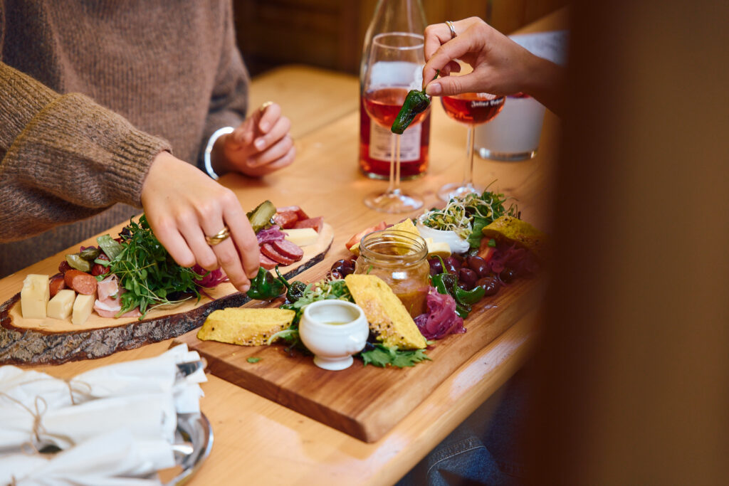 Nahaufnahme eines Holztisches mit reichlich gef&uuml;llten Vesperplatten. Auf einem Holzbrett und einer Baumscheibe liegen Schinken, K&auml;se, Wurst, Brot, Oliven, frische Kr&auml;uter und Dips. Im Hintergrund stehen eine Flasche Ros&eacute;wein sowie zwei gef&uuml;llte Weingl&auml;ser, von denen eine Hand eines Gastes gerade eines greift.