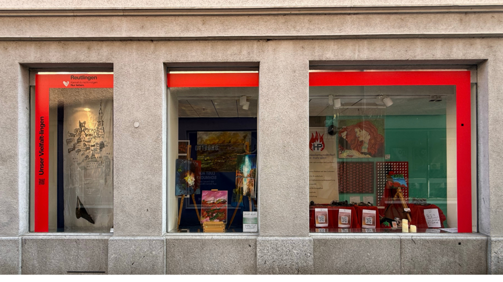 Außenansicht eines Gebäudes mit drei großen Schaufenstern einer Galerie oder Kunstausstellung. Die Fenster sind mit leuchtend roten Rahmen eingefasst. Links steht in weißer Schrift auf rotem Grund „Unser Vielfalt-lingen“ und „Reutlingen“. In den Fenstern sind verschiedene Kunstwerke, Gemälde auf Staffeleien, Infoplakate und QR-Codes ausgestellt.