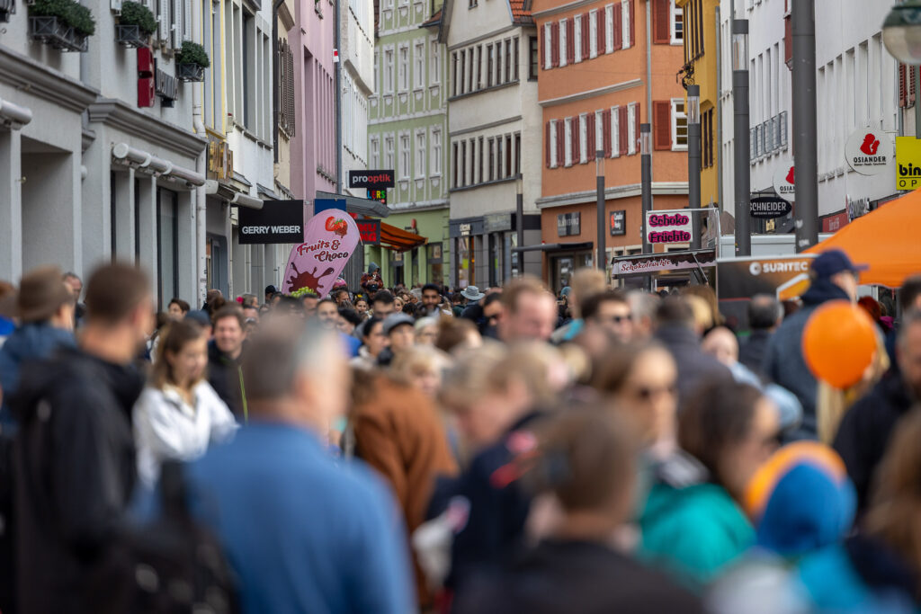 Eine Aufnahme einer belebten Fu&szlig;g&auml;ngerzone oder Einkaufsstra&szlig;e in einer historischen Stadt, gef&uuml;llt mit einer dichten Menschenmenge. Im Hintergrund sind mehrst&ouml;ckige Geb&auml;ude mit traditioneller Architektur, Schaufenstern und verschiedenen Gesch&auml;ftsschildern (unter anderem &bdquo;Gerry Weber&ldquo; und &bdquo;prooptik&ldquo;) zu sehen. Einige Personen im Vordergrund sind unscharf, w&auml;hrend die Menge sich die Stra&szlig;e entlangbewegt. Auf der rechten Seite ist ein oranges Schild oder ein Luftballon zu erkennen.
