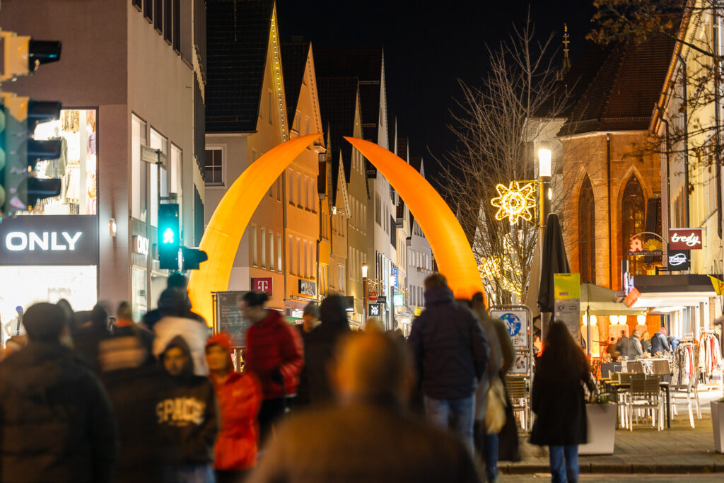 Belebte Fußgängerzone am Abend mit weihnachtlicher Beleuchtung, Passanten und zwei großen, orangefarben leuchtenden Bögen.