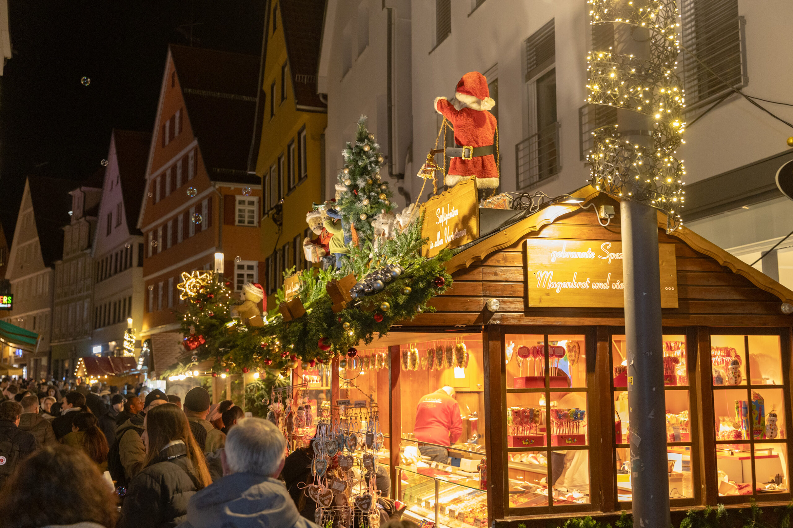 Belebte Fu&szlig;g&auml;ngerzone am Abend mit weihnachtlicher Beleuchtung, Passanten und zwei gro&szlig;en, orangefarben leuchtenden B&ouml;gen.