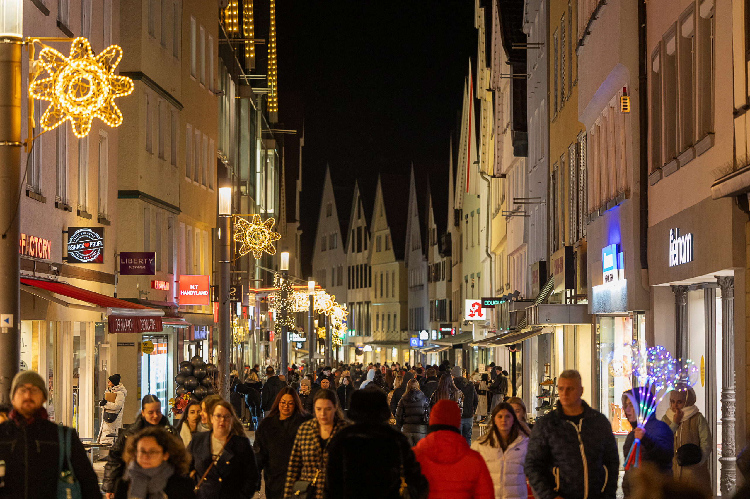 Belebte Fu&szlig;g&auml;ngerzone am Abend mit weihnachtlicher Beleuchtung, Passanten und zwei gro&szlig;en, orangefarben leuchtenden B&ouml;gen.